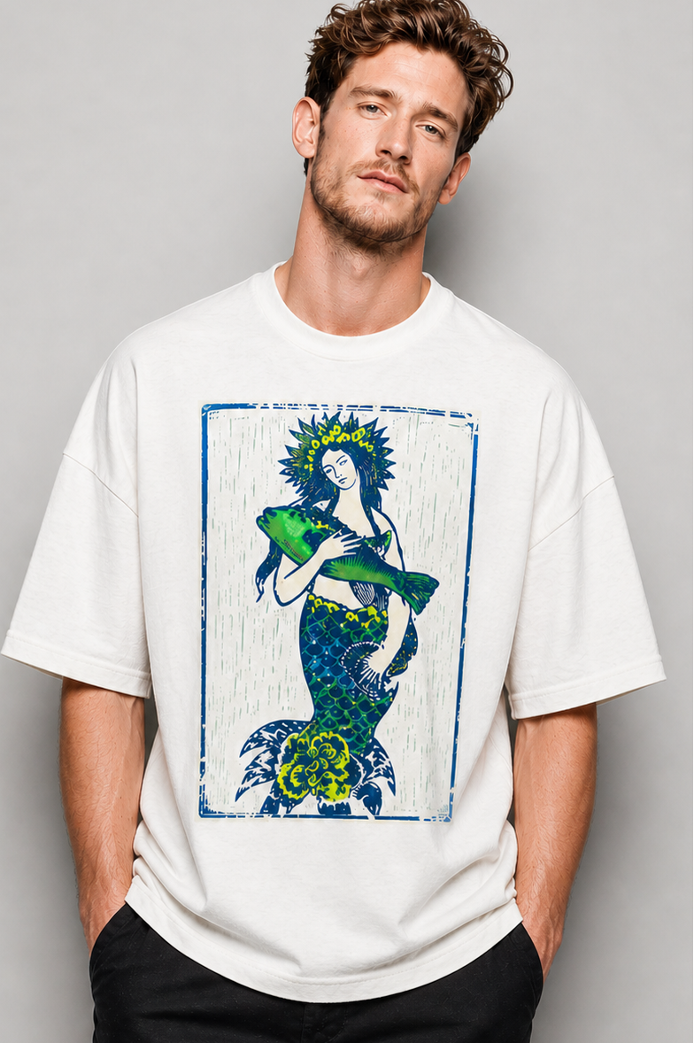 Man wearing a white t-shirt with a graphic design of a mermaid holding a fish, standing against a gray background.