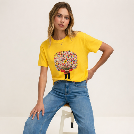 Woman wearing a yellow t-shirt with a colorful design and blue jeans, sitting on a white stool.