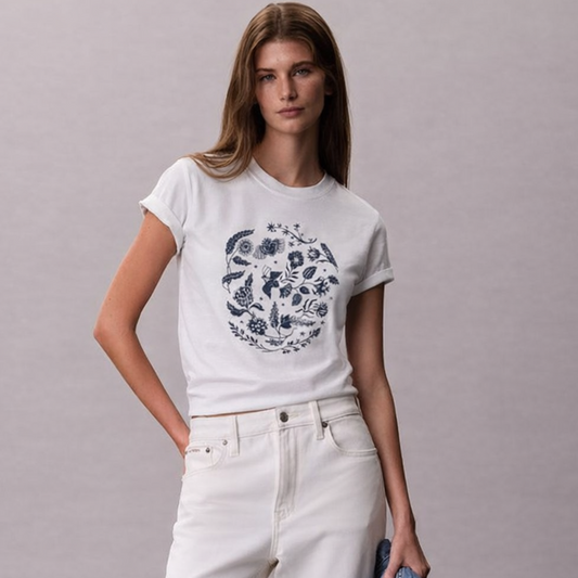 Woman wearing a white t-shirt with a graphic design and white pants, holding a denim jacket against a plain background.