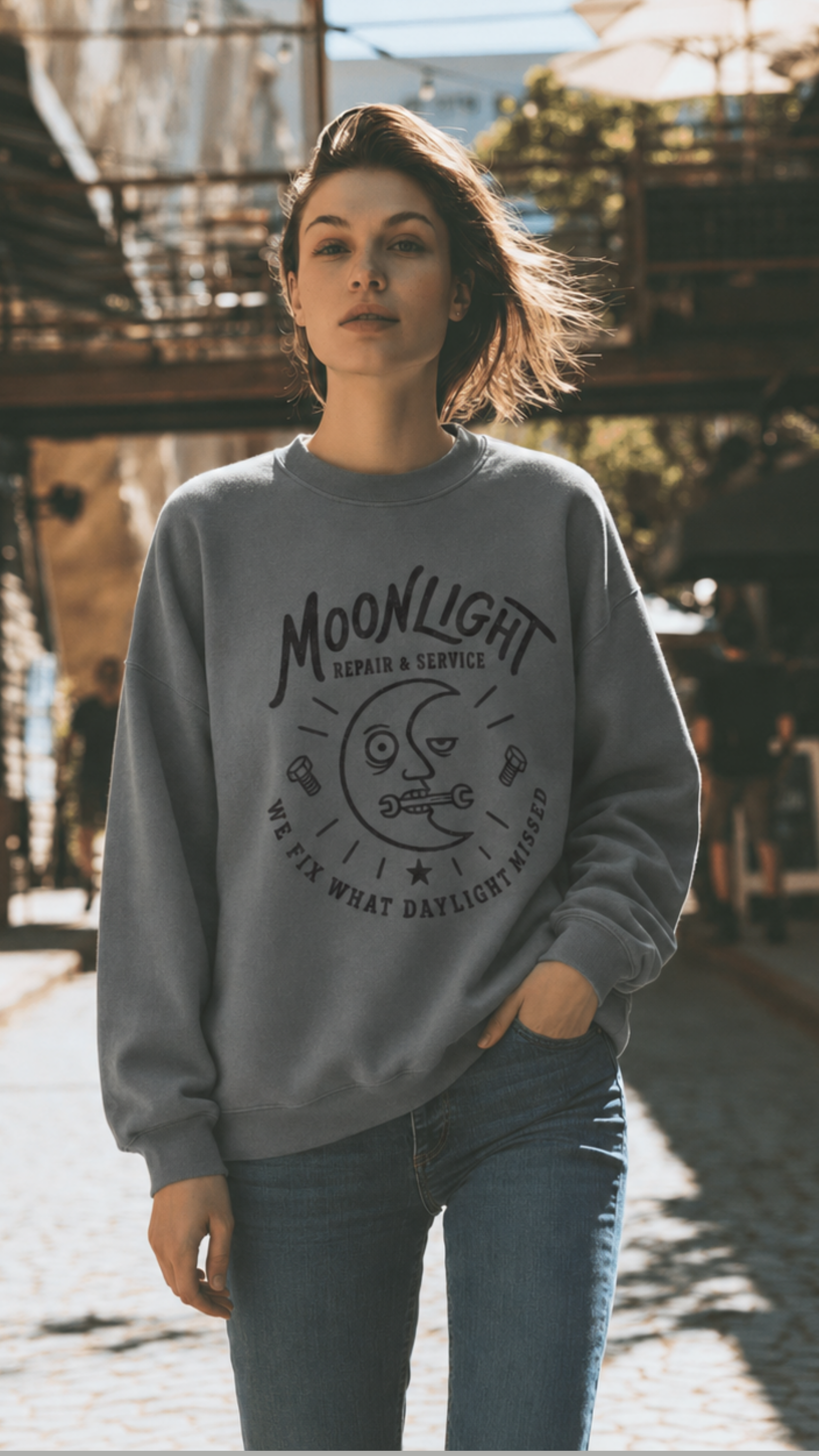 A woman stands on a sunlit cobblestone street wearing a gray crewneck sweatshirt with a black graphic that reads 'MOONLIGHT' and 'WE ARE WHAT SATELLITES ARE'.